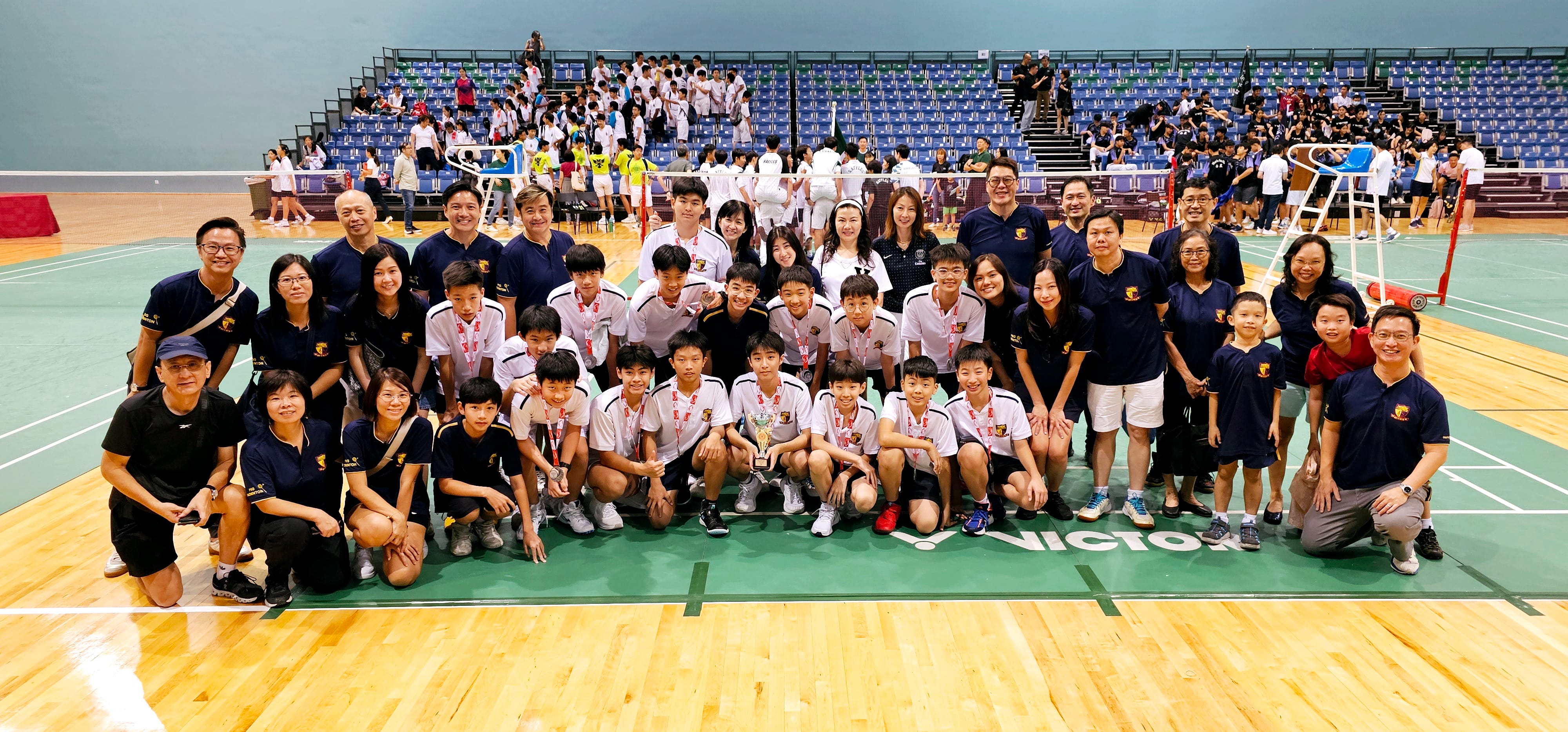 A large badminton squad poses together on a wooden court in a sports hall, arranged in rows with some players kneeling in front.
